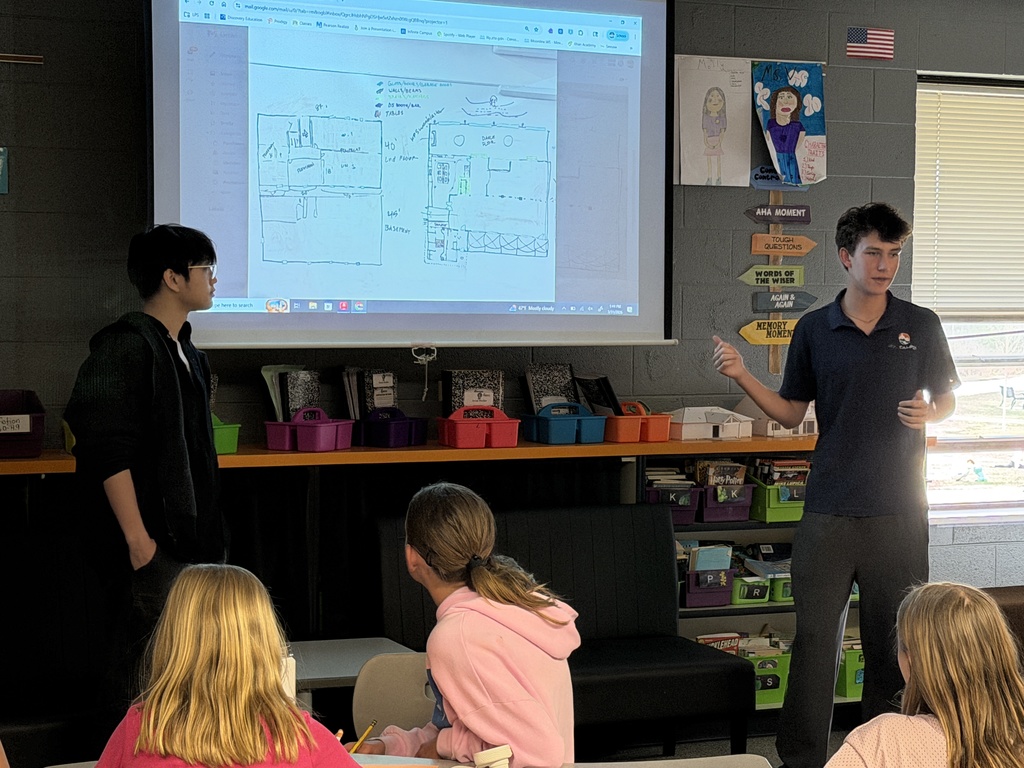Two high school students stand at the front of a classroom presenting to peers. One student gestures toward a projected screen displaying a hand-drawn floor plan, while the other stands beside it. Several younger students sit at desks facing them. The classroom includes shelves with colorful bins, posters on the wall, and natural light from a window.