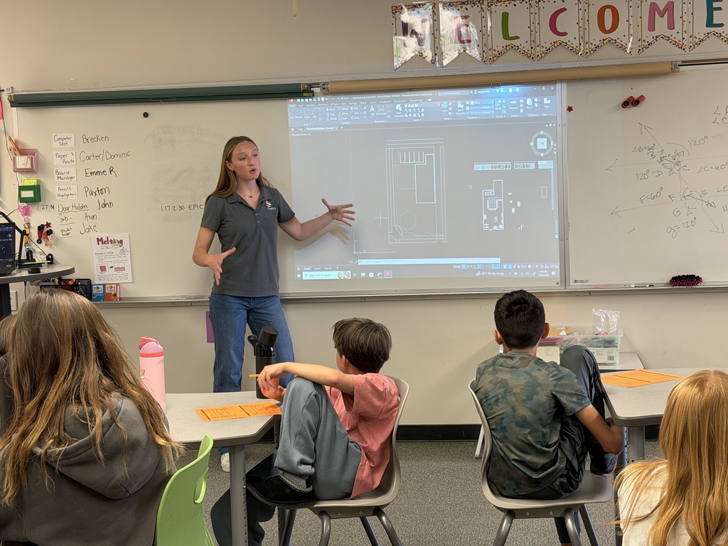 A female instructor stands at the front of a classroom, gesturing with both hands while presenting a digital floor plan projected onto a whiteboard. Elementary-aged students sit at desks facing her, some holding pencils and worksheets. A “WELCOME” banner hangs above the board, and handwritten notes and diagrams are visible on the whiteboard.