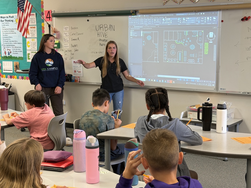 Two female presenters stand at the front of a classroom leading a lesson while a floor plan design is projected on the whiteboard behind them. One gestures toward the screen while speaking. Elementary students sit at desks with worksheets and water bottles, facing the presenters. The board includes the heading “URBAN HIVE” and classroom posters and schedules are visible.