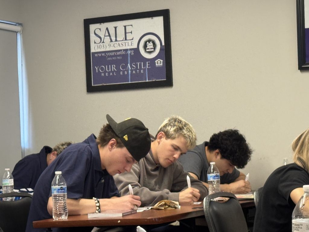 A closer view of three high school students seated at a table, concentrating as they write on worksheets. One student wears a backward cap, and water bottles and papers are on the table. A framed poster reading “SALE Your Castle Real Estate” hangs on the wall behind them.