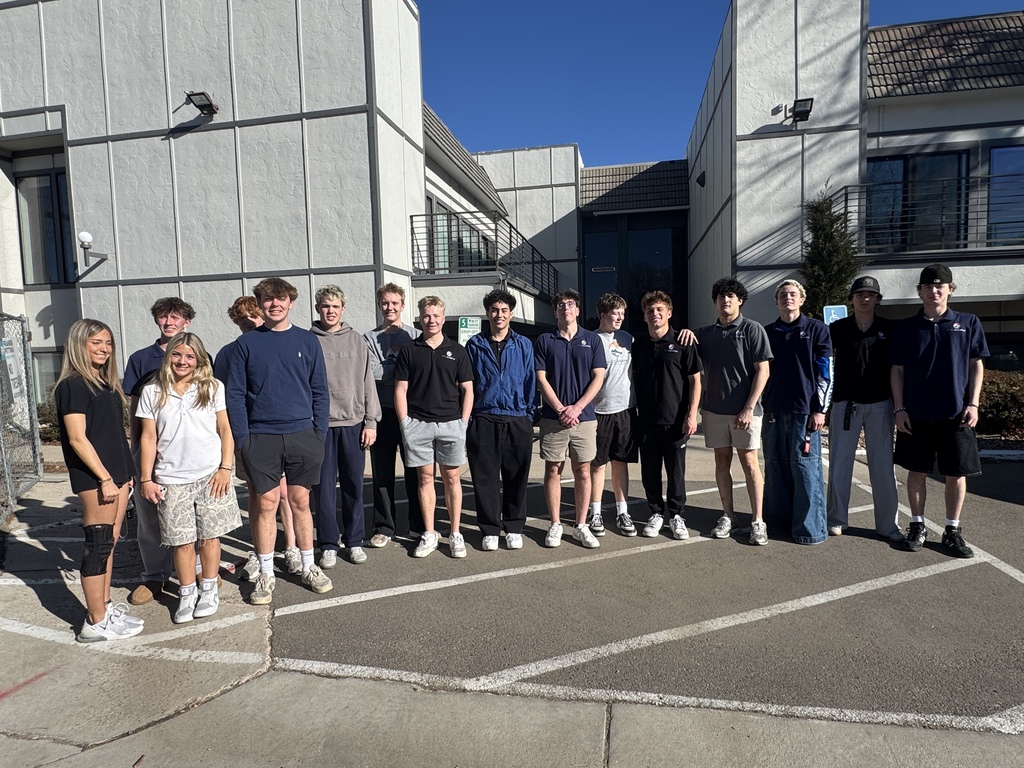 A group of high school students of diverse backgrounds stands outdoors in a parking lot in front of a modern school building, smiling at the camera. Students wear a mix of EPIC Campus polos, hoodies, and casual clothing such as shorts and sneakers. Bright sunlight casts strong shadows, and the building’s light-colored exterior, windows, and balcony railings are visible behind them.