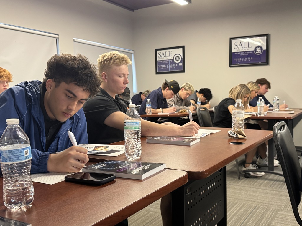 High school students sit at classroom tables writing on worksheets, focused and quiet. Water bottles and booklets are on the desks. The room has framed posters on the wall and window shades pulled down, creating a classroom or training setting.