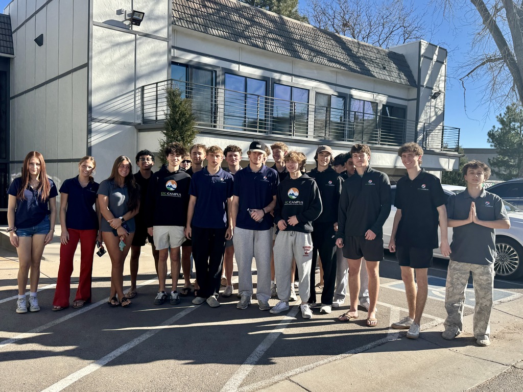 A large group of high school students of diverse backgrounds stand outdoors in front of a modern school building, smiling at the camera. Most wear EPIC Campus polos or hoodies, while a few wear casual outfits. The group stands in a parking lot under bright sunlight, with shadows cast on the ground and the building’s windows and balcony visible behind them.