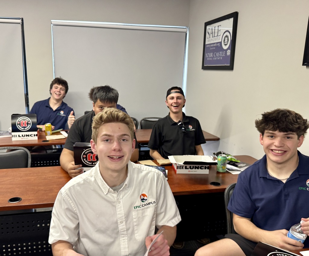 A group of high school students sits in a classroom at tables, smiling toward the camera while holding boxed lunches. Several wear EPIC Campus polos, and one student gives a thumbs-up. The room includes framed wall posters reading “SALE Your Castle Real Estate,” and window shades partially drawn behind them.