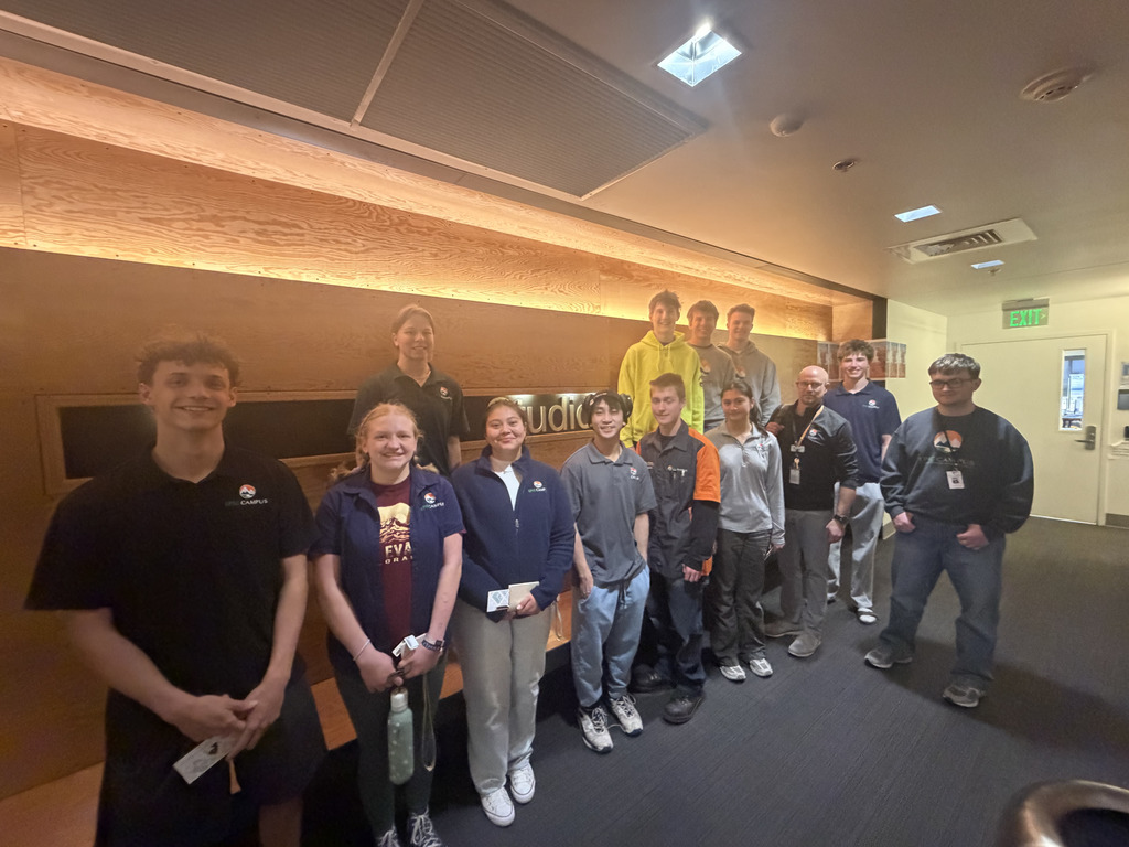 A group of high school students and a staff member stand together indoors in a modern office space with warm wood paneling and a sign on the wall. Students wear EPIC Campus apparel and casual clothing, smiling at the camera while holding small items or notebooks.