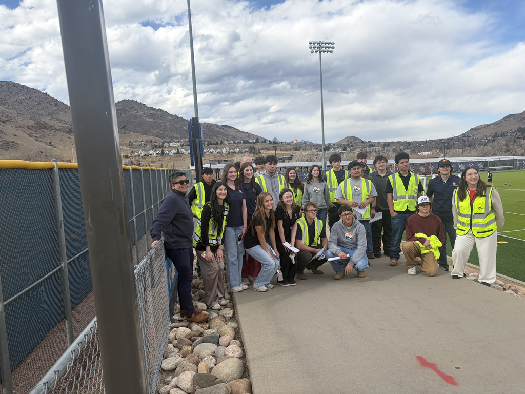 A group of high school students and an adult stand outdoors near a fenced athletic field, wearing bright safety vests and casual clothing. Some hold papers as they pose together on a paved walkway. Stadium lights, mountains, and a cloudy sky are visible in the background.