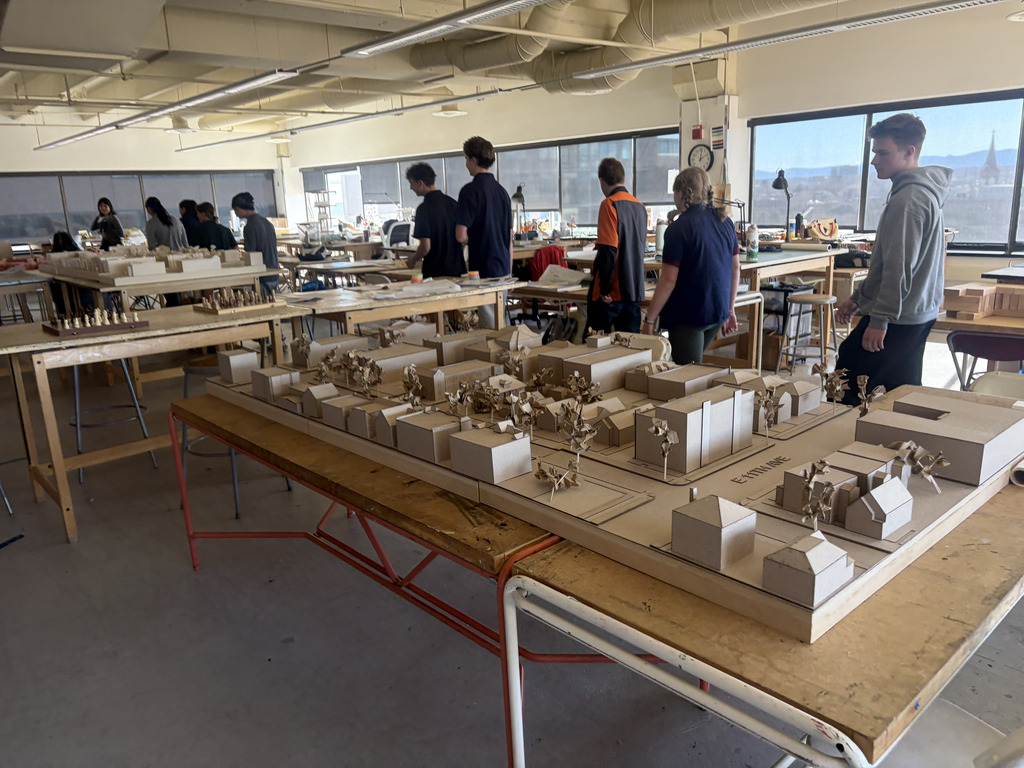 Students walk through a bright classroom or studio filled with tables displaying detailed architectural models made of cardboard. Large windows line the room, and students in casual clothing observe the models and workspace.