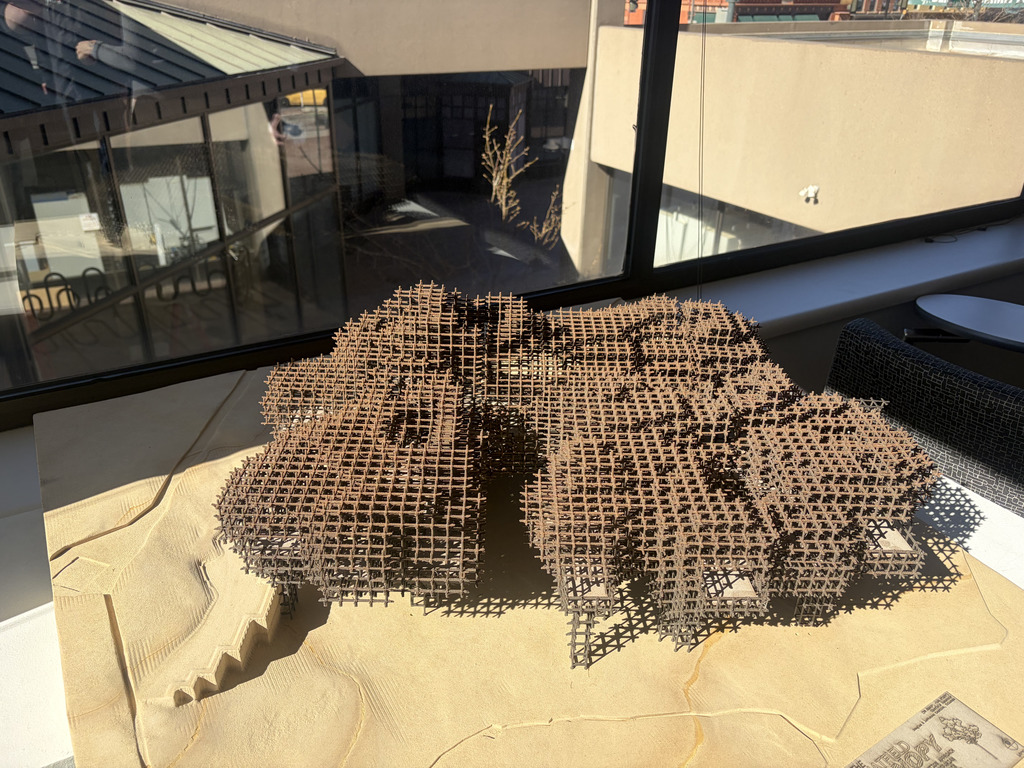 A close-up of an intricate architectural model made of thin wooden lattice pieces forming a curved, layered structure on a base. The model sits on a table near large windows with sunlight casting shadows across the design and the surrounding indoor space.