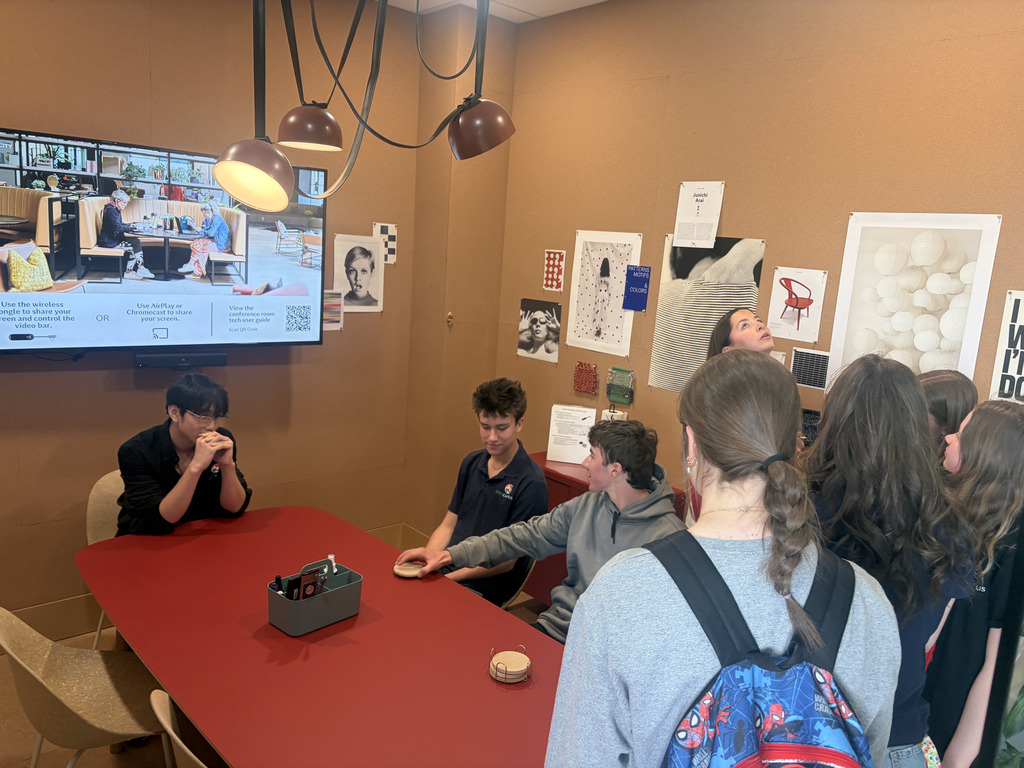 Students gather around a table in a small meeting room with orange walls, watching as two students sit at the table and interact with objects. A wall-mounted screen displays instructions about screen sharing, and design posters and sample materials are pinned to the walls.
