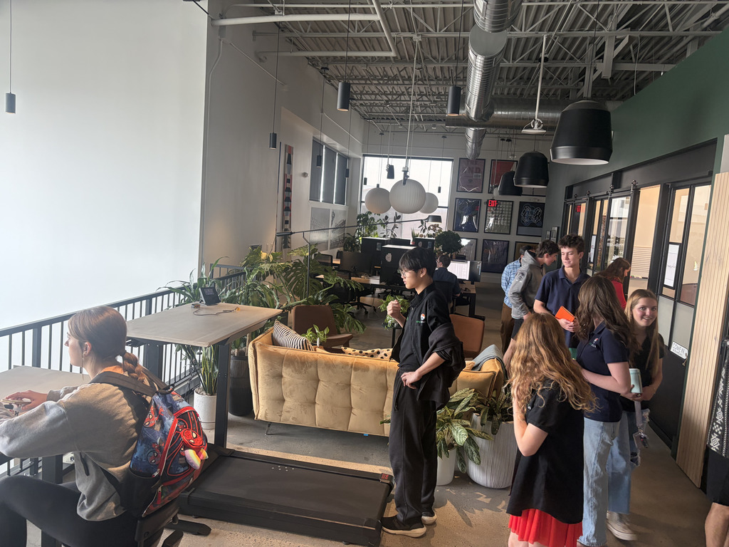 Students explore a modern office space with high ceilings, exposed ductwork, and large windows. One student sits at a desk working on a computer while others stand nearby talking and observing. The space includes a couch, indoor plants, hanging lights, and glass-walled rooms.