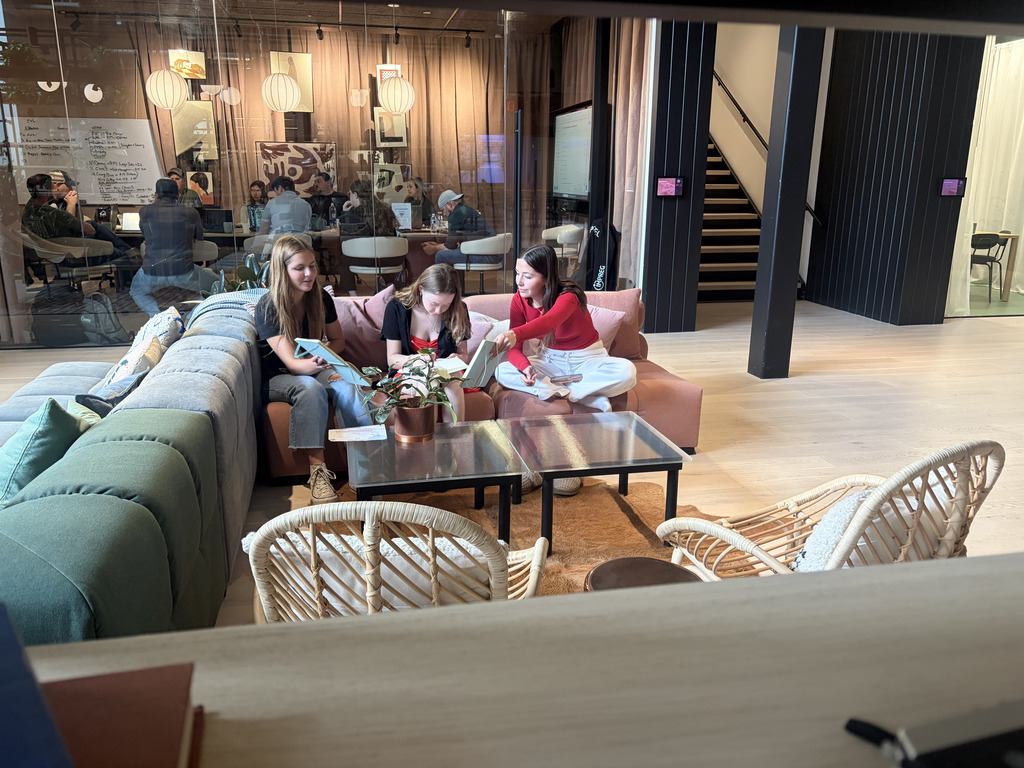 Three high school students sit on a couch in a stylish lounge area, looking at tablets and discussing something together. A small table with a plant sits in front of them, and behind them a glass-walled conference room shows people meeting inside.