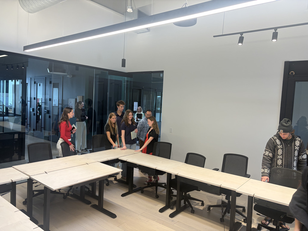 A group of high school students stands around a conference table in a modern meeting room with glass walls and overhead lighting. Some hold notebooks while listening and talking, as one student adjusts a chair. The room has multiple rolling chairs and a clean, minimalist design.