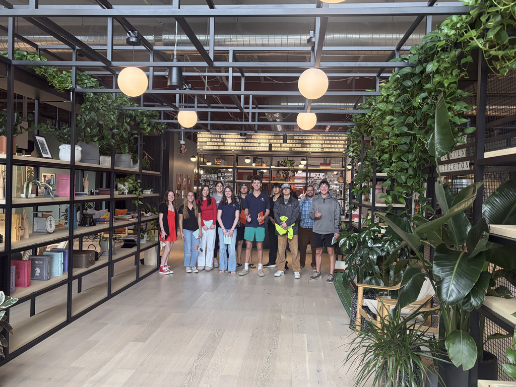 A group of high school students stands together inside a modern, plant-filled workspace with open shelving and hanging globe lights. Students wear a mix of EPIC Campus and casual clothing, smiling at the camera in a bright, collaborative environment.