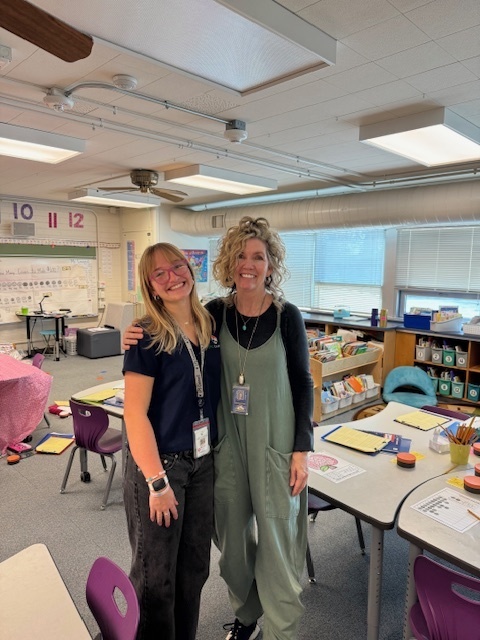 Two educators stand together in a classroom, smiling with their arms around each other. The room includes desks, colorful chairs, bookshelves, and educational materials, creating a welcoming learning environment.