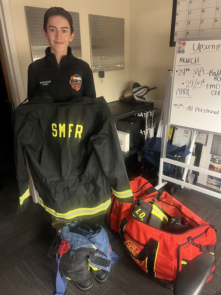 A student intern stands indoors holding a black firefighter jacket with bright yellow reflective stripes and large “SMFR” lettering on the back. Nearby are firefighter gear bags and equipment. A whiteboard behind them lists upcoming dates and events.