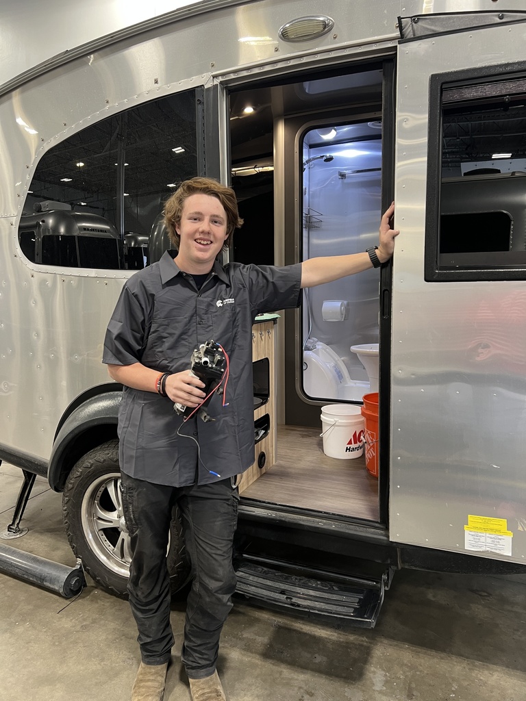 A student stands at the open door of a small trailer or RV, holding a mechanical component with wires. Inside the trailer, a compact bathroom and cabinetry are visible. The student wears work clothes and smiles at the camera.