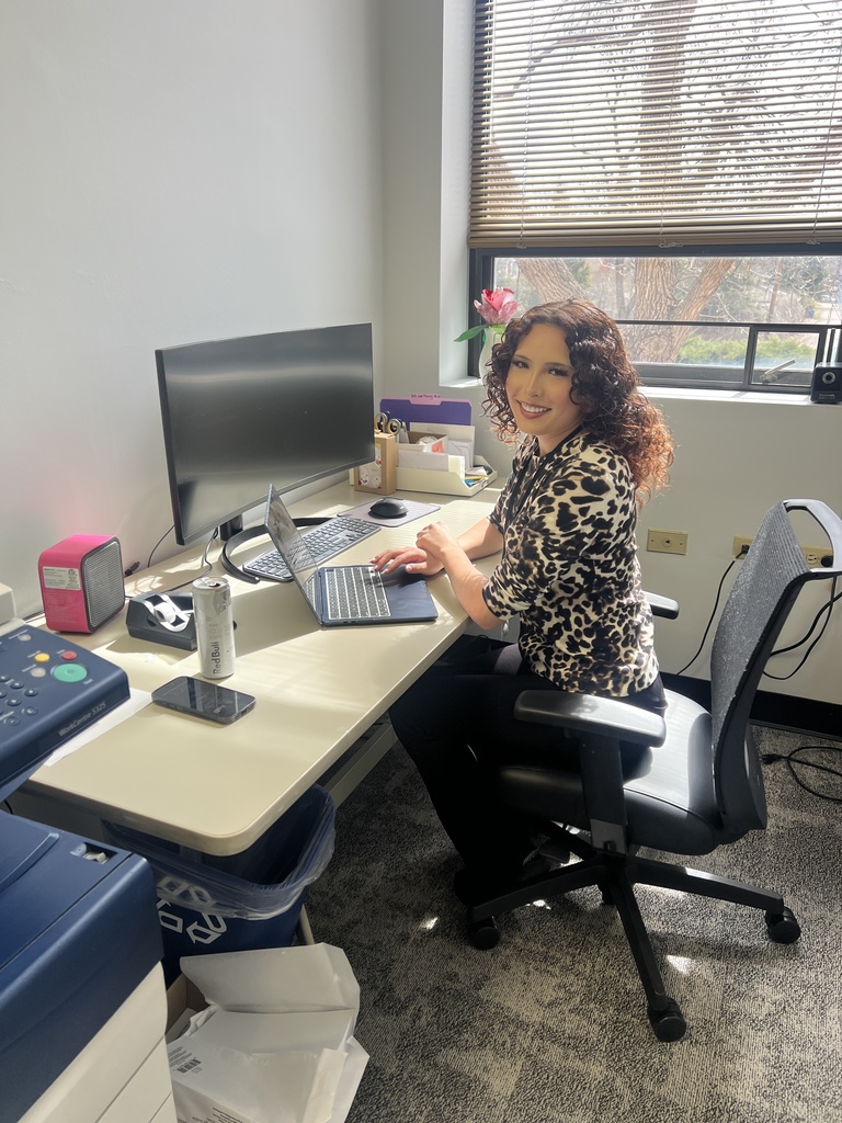 A staff member with curly hair sits at a desk in an office, smiling while typing on a laptop. Sunlight streams through a window with blinds behind her. The desk holds a monitor, keyboard, phone, drink can, and office supplies.