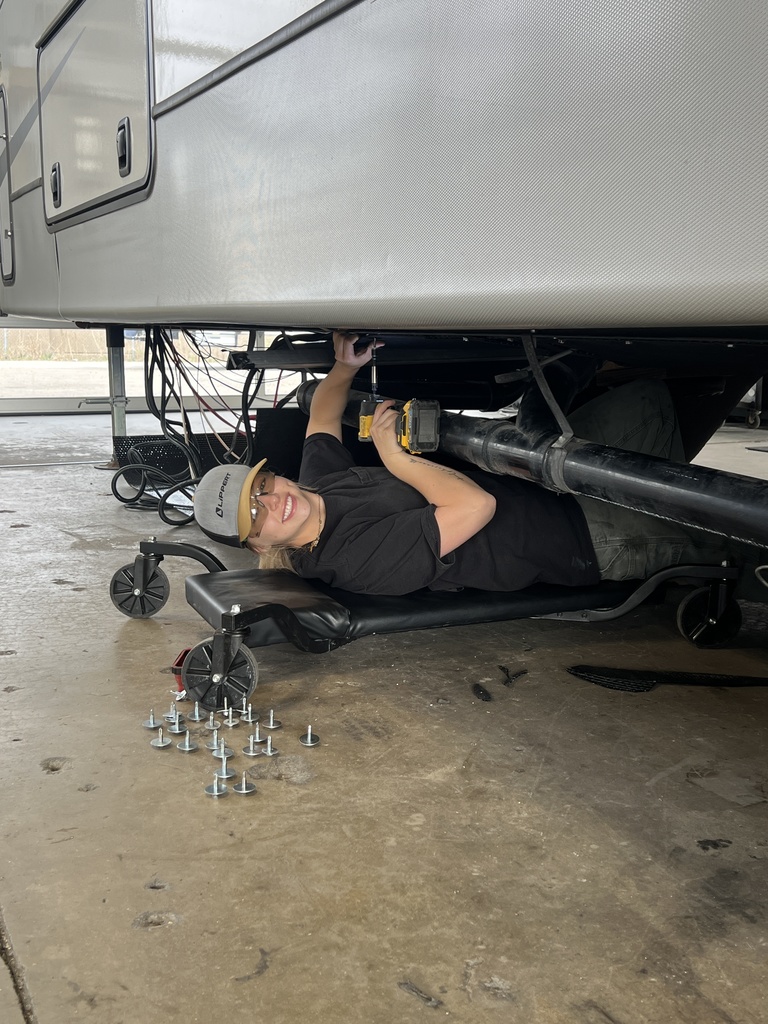 A student lies on a rolling mechanic’s creeper under a large vehicle, smiling while using a power tool to work underneath. Several bolts are arranged on the floor nearby, and the setting appears to be an auto or technical workshop.