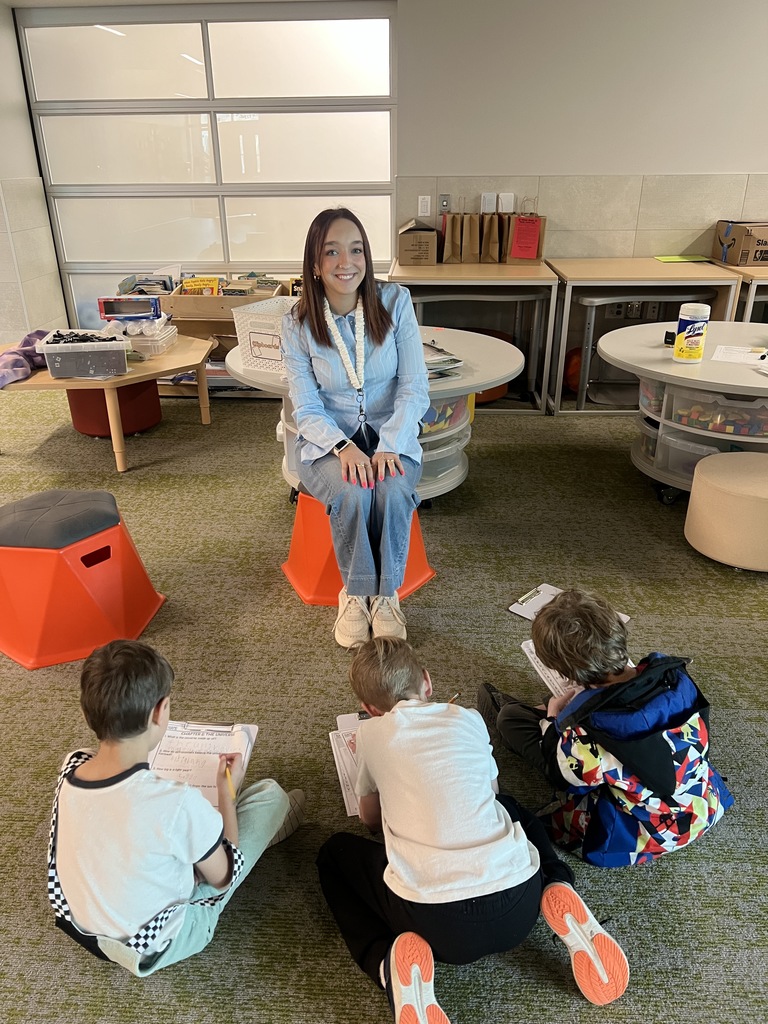 An elementary classroom scene where a young teacher sits on a small orange stool, smiling at three students seated on the floor in front of her. The students, working on papers with pencils, face the teacher. The room includes low tables, bins of classroom materials, and a soft carpeted area.