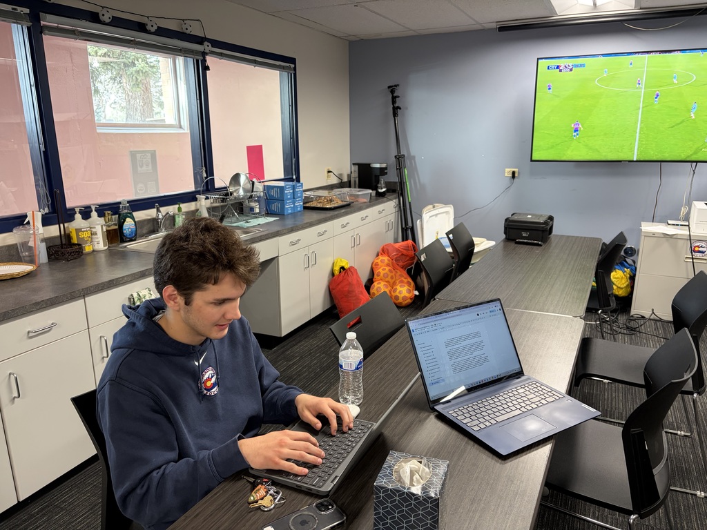 A student sits at a long table in a classroom or workroom, focused on typing on a laptop. Another laptop, a water bottle, keys, and a tissue box are on the table. In the background are cabinets, a sink, and a wall-mounted TV displaying a soccer game.