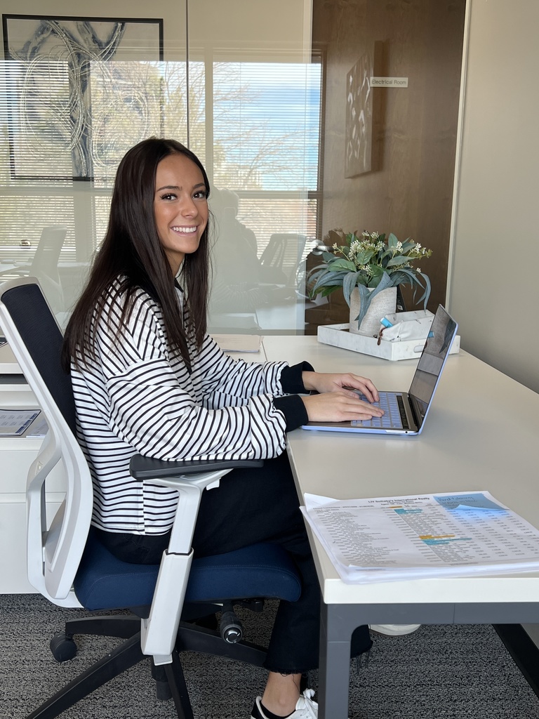A staff member sits at a desk in a glass-walled office, smiling while working on a laptop. Papers are spread on the desk, and a sign on the door reads “Electrical Room.” A small plant and office items decorate the space.