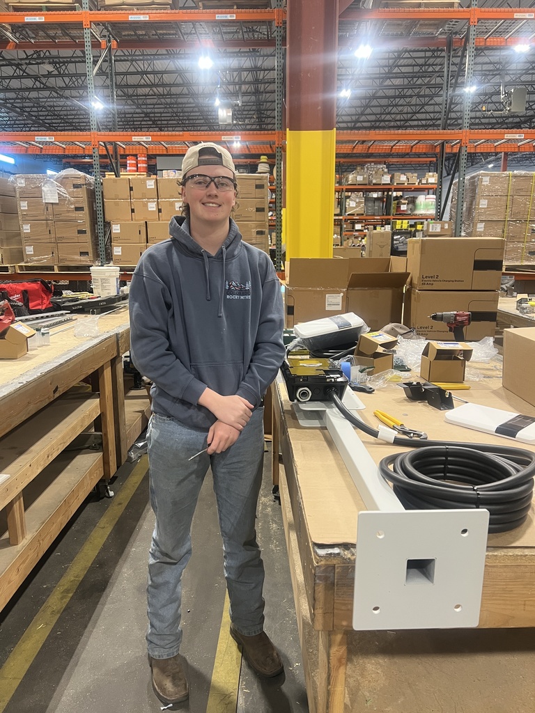 A high school–aged student wearing safety glasses, a backwards cap, a gray hoodie, jeans, and work boots stands in a large warehouse. They smile while holding a small tool beside a workbench covered with electrical components, cables, and boxes. Tall industrial shelving filled with labeled boxes and bright overhead lights fills the background.