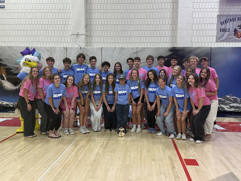 A large group of students in pink and blue Heritage Wish Week shirts pose together with an eagle mascot in a gymnasium with special guest wish kid Colton.