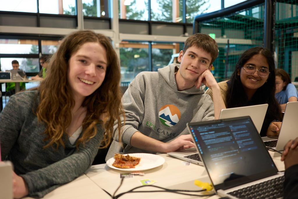 Three high school students sit together at a table with laptops in a bright, modern learning space. A student in the center wears a gray EPIC Campus hoodie, and a slice of pizza sits on a plate in front of the group.