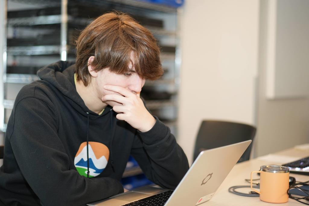 A high school student in a black EPIC Campus hoodie looks thoughtfully at a laptop while resting a hand near their face. A mug and computer equipment sit on the table beside them.