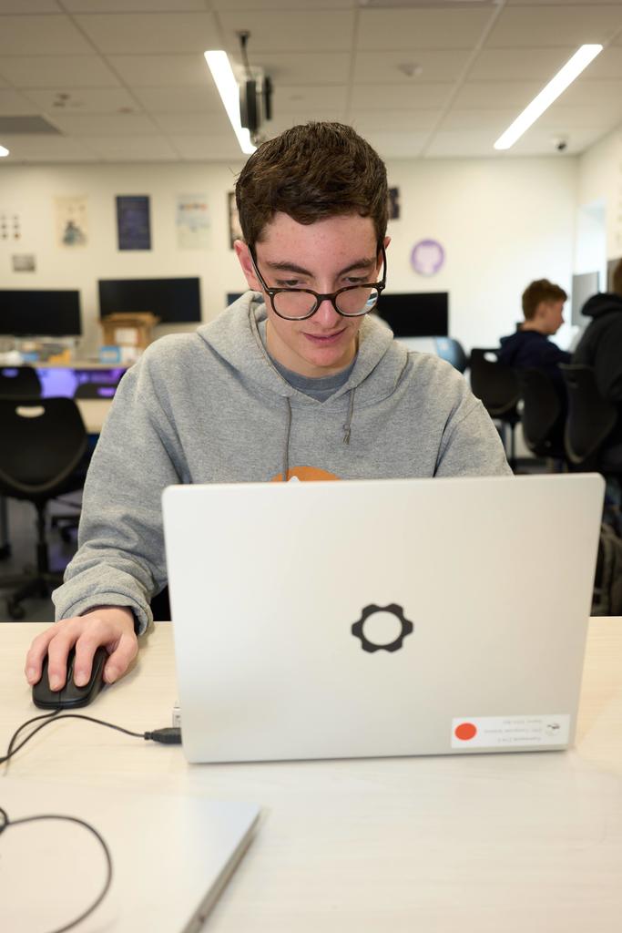 A high school student wearing glasses and a gray EPIC Campus hoodie works at a laptop with a mouse in a classroom computer lab. Other students and computer monitors are visible, softly blurred in the background.