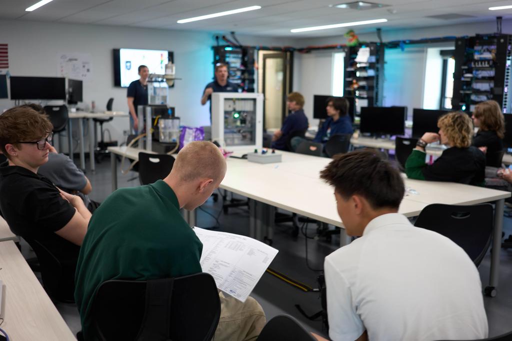 Students sit around tables in a classroom lab while an instructor stands at the front explaining a technical setup. In the foreground, two students review a printed worksheet, while computer stations and equipment racks line the room.