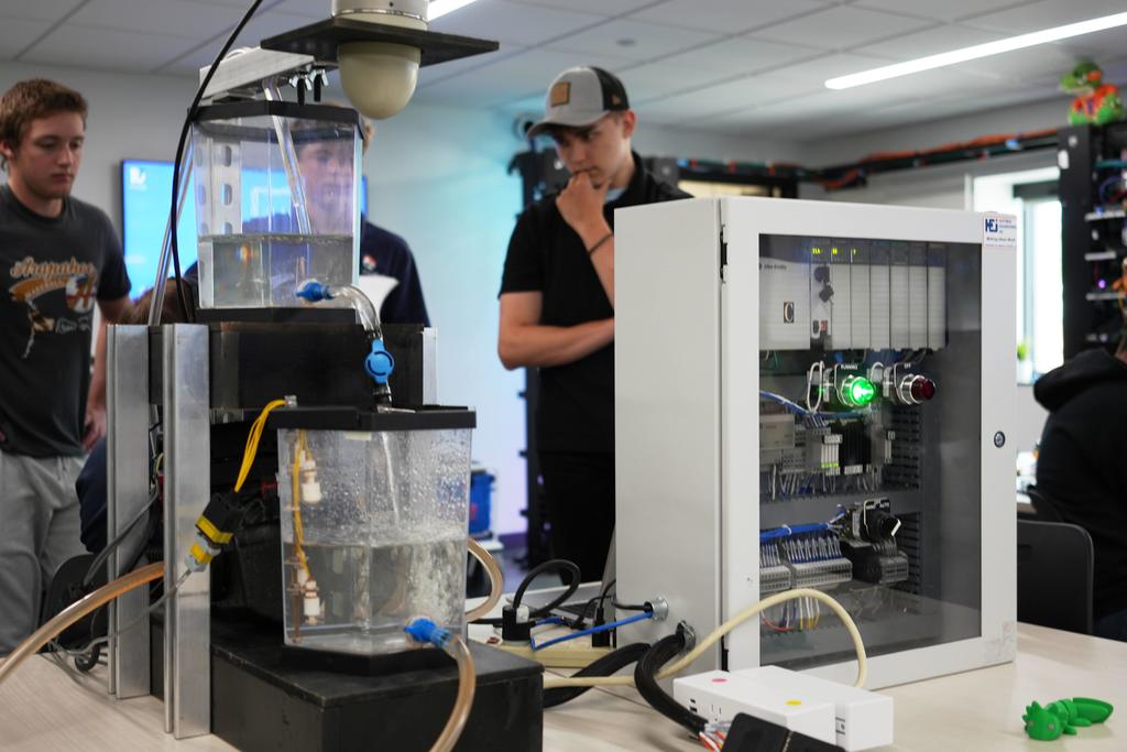 Close-up of a classroom engineering setup with clear water tanks, tubing, and sensors connected to a control panel with a glowing green indicator light. Several high school students stand nearby observing the system, one wearing a cap and resting a hand on their chin.