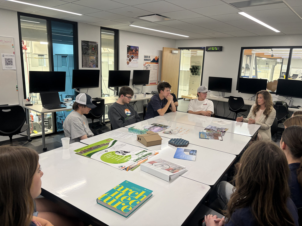 A wider view of the same classroom shows a diverse group of high school students—boys and girls—seated around connected white tables while a female adult speaker leads a discussion. Students wear casual clothing, including T-shirts and baseball caps, and appear engaged, some leaning forward or resting their chins on their hands. The tables are covered with brochures, booklets (including one titled “architectural guide”), a small cardboard building model, and design samples. Multiple computer monitors line the walls, and large interior windows reveal a workshop or lab space beyond. Posters and a digital clock are visible on the walls, reinforcing the classroom’s academic and career-focused environment.