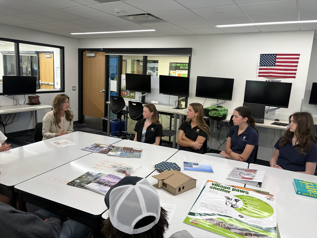 A group of five high school students—mostly girls wearing matching dark polo shirts—sit around white tables in a modern classroom, listening to a female adult speaker in a light blazer. The students appear attentive, facing the speaker with hands folded or resting on the table. On the tables are brochures, printed materials, a small cardboard architectural model, and a hexagonal tile sample. Behind them are computer monitors along the wall, a glass door, and a U.S. flag mounted on the wall. The room is bright with overhead lighting and has a clean, professional lab or classroom setting.