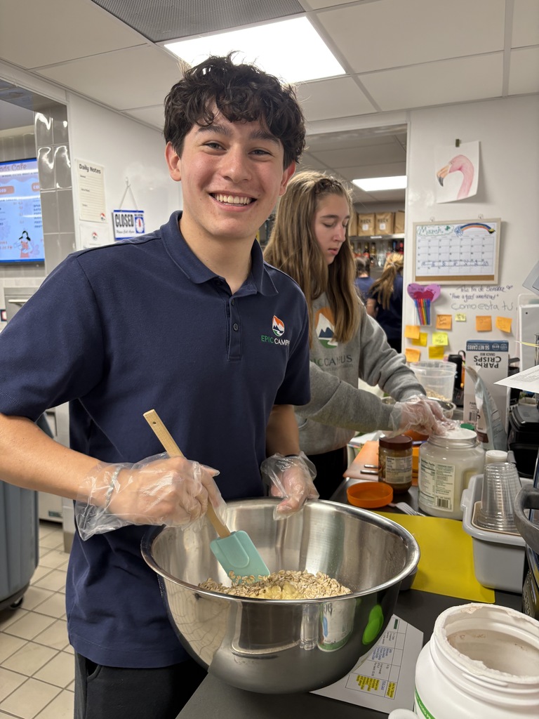 A high school boy smiles while mixing ingredients in a large metal bowl using a spatula. Another student works behind him at the counter. Both wear gloves, and the workspace includes containers, measuring tools, and a colorful bulletin area with notes and a calendar in the background.