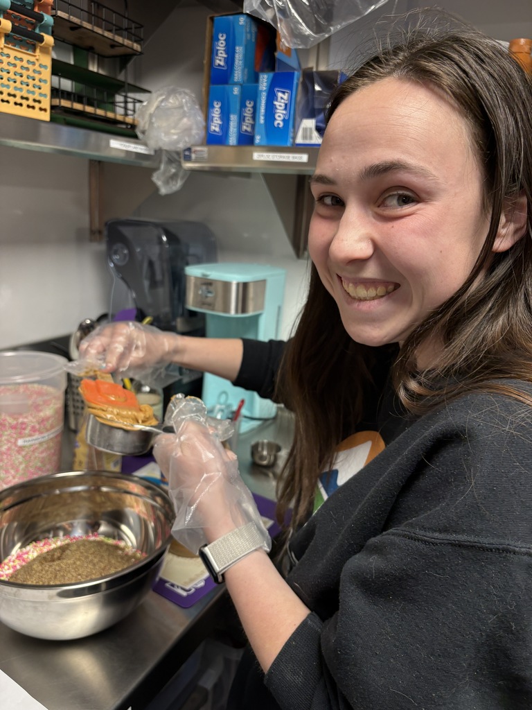 A close-up of a high school girl smiling while scooping ingredients onto a snack mixture. She wears gloves and stands at a counter with bowls of oats and sprinkles, surrounded by kitchen supplies and storage shelves.
