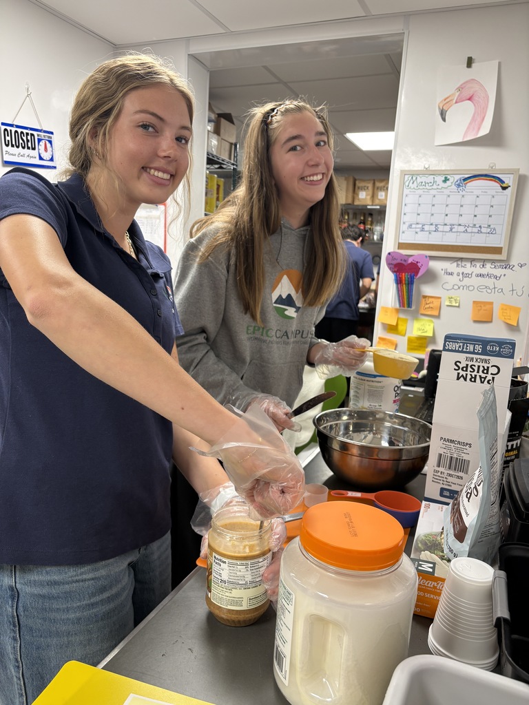 Two high school girls smile at the camera while preparing food in a classroom kitchen area. Both wear gloves and are mixing ingredients at a counter with bowls, measuring cups, and containers. Behind them are shelves, a calendar labeled “March,” colorful sticky notes, and a sign that reads “Closed.”