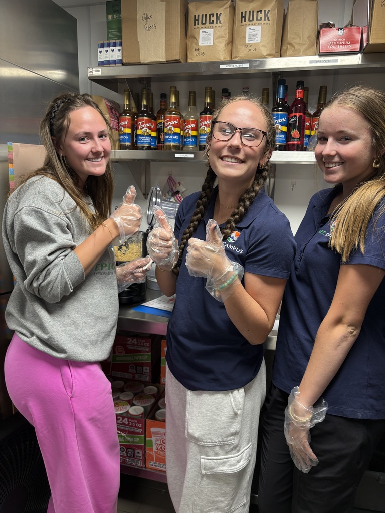 Three high school girls stand in a small kitchen workspace, smiling and giving thumbs up while wearing gloves. Behind them are shelves stocked with coffee syrups, filters, and labeled containers, suggesting a student-run café or food prep area.