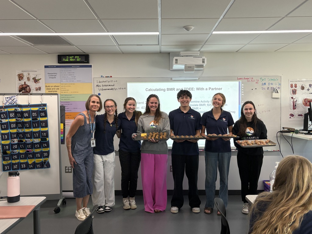 A teacher stands with a group of seven high school students in a classroom, all smiling and facing the camera. Several students hold trays of homemade snack bites. A projection screen behind them reads “Calculating BMR and TDEE: With a Partner,” and classroom posters, a wall clock, and a numbered pocket organizer are visible. The students wear a mix of navy school polos and casual clothing, representing a diverse group.