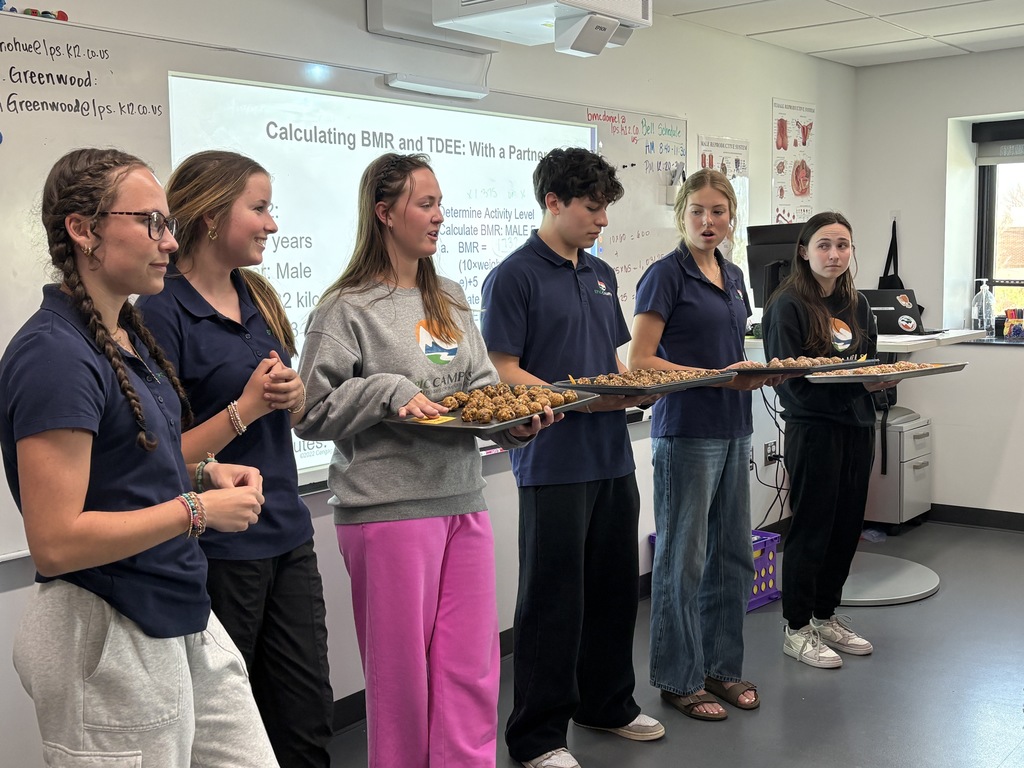 A line of high school students stands at the front of a classroom presenting trays of homemade snack bites while speaking. A projected slide behind them reads “Calculating BMR and TDEE: With a Partner.” The students wear navy polos and casual attire, and appear engaged in explaining their work to classmates.