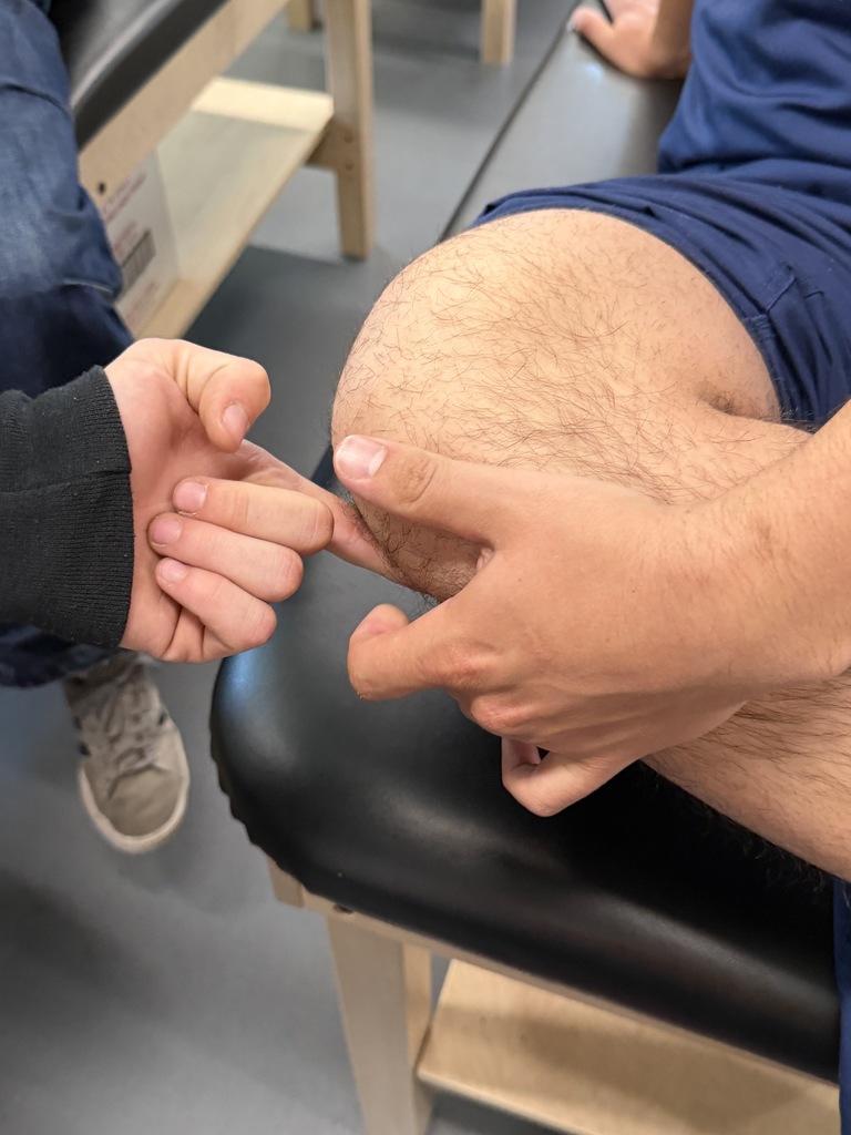 A person sits on a black bench, wearing blue shorts. Another individual touches their knee with a finger.