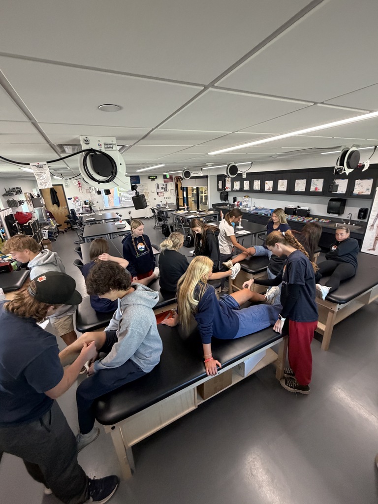 A room with students sitting on tables and performing massages. A person is standing near them.