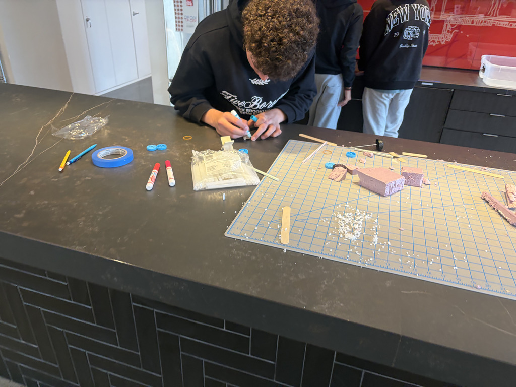 Person seated at a countertop, crafting with various art tools and materials. A gridded sheet is on the counter.