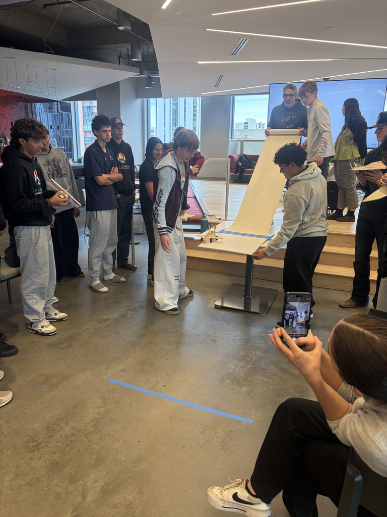 Several people observe a group of individuals rolling a large white sheet on a stand. One person films with a phone.
