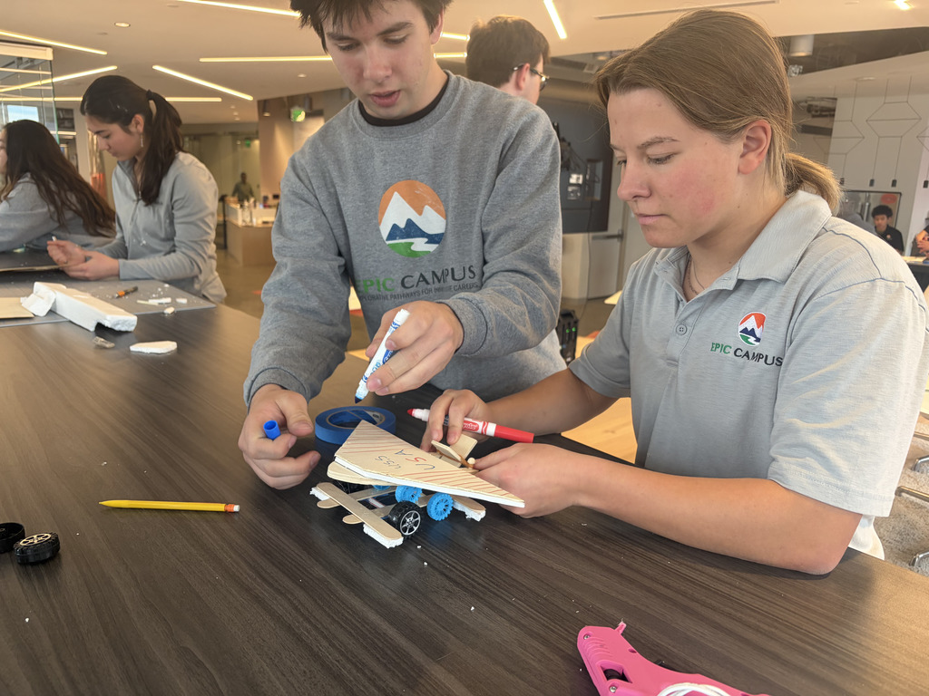 A man and a woman decorate a miniature airplane model with brushes and colored pencils at a table.