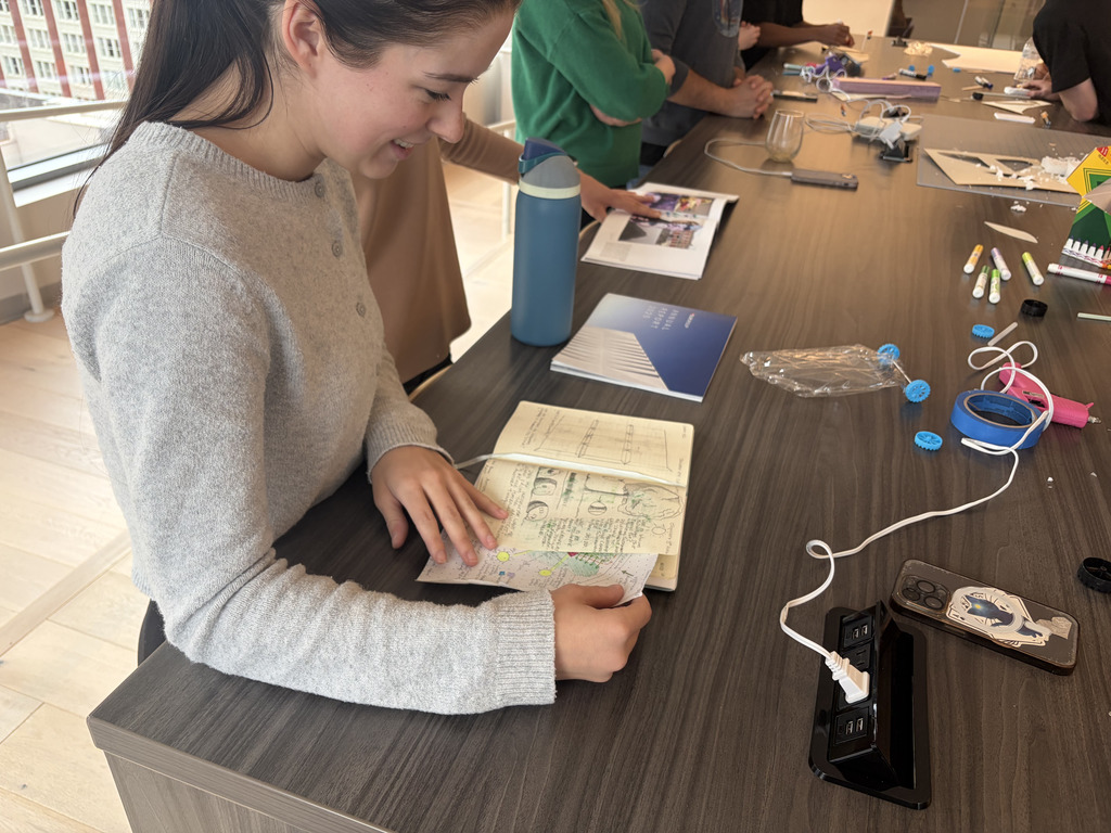 Person seated at a desk, holding a book with hands, with various objects like a phone and a bottle.