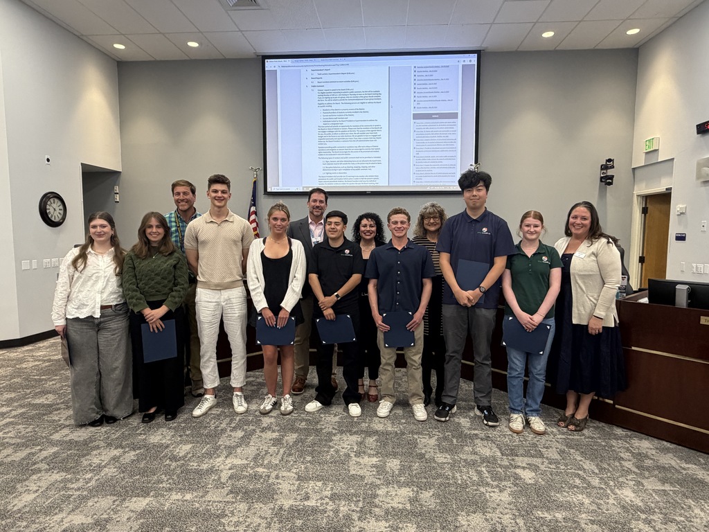 A group of students in a meeting room, holding certificates, stands before a screen displaying text.