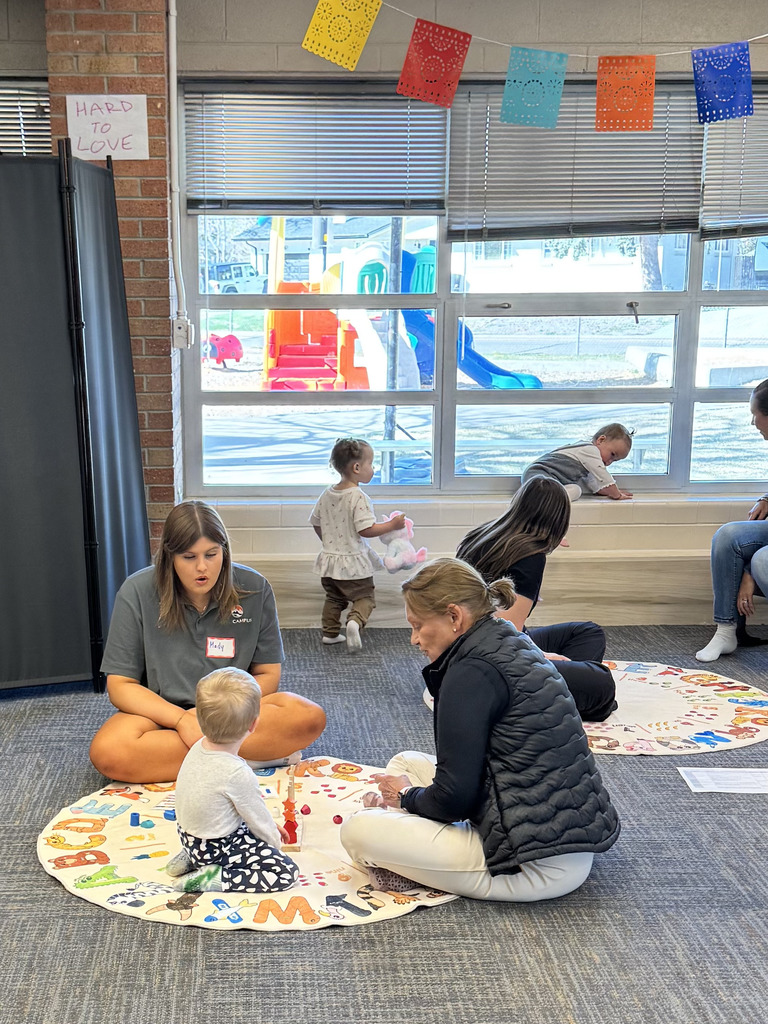 A room with adults and children, one group sitting around a mat with letters. A playground is visible through the windows.
