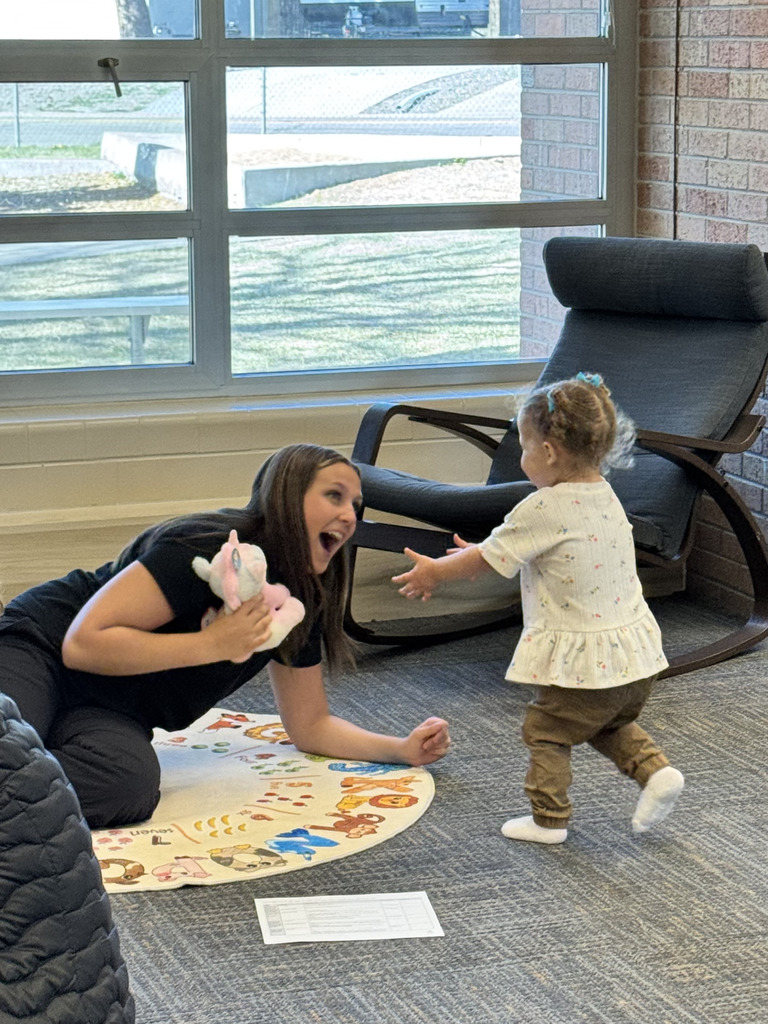 A woman crouches, holding a stuffed animal, as a toddler runs towards her, both smiling. Behind them, a black lounge chair and a window.