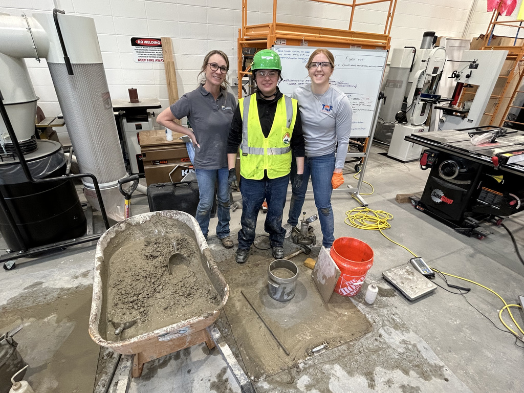 Three people stand in a room with construction tools. One wears a green helmet and vest, another wears a white shirt, and the third wears a gray shirt.